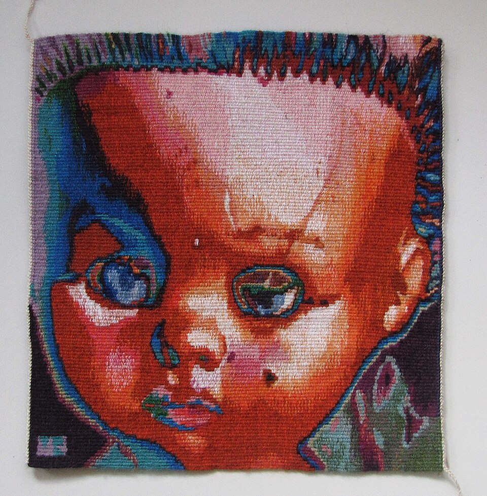 A small woven tapestry depicting a doll’s face in red, blue, green and pale pink threads.