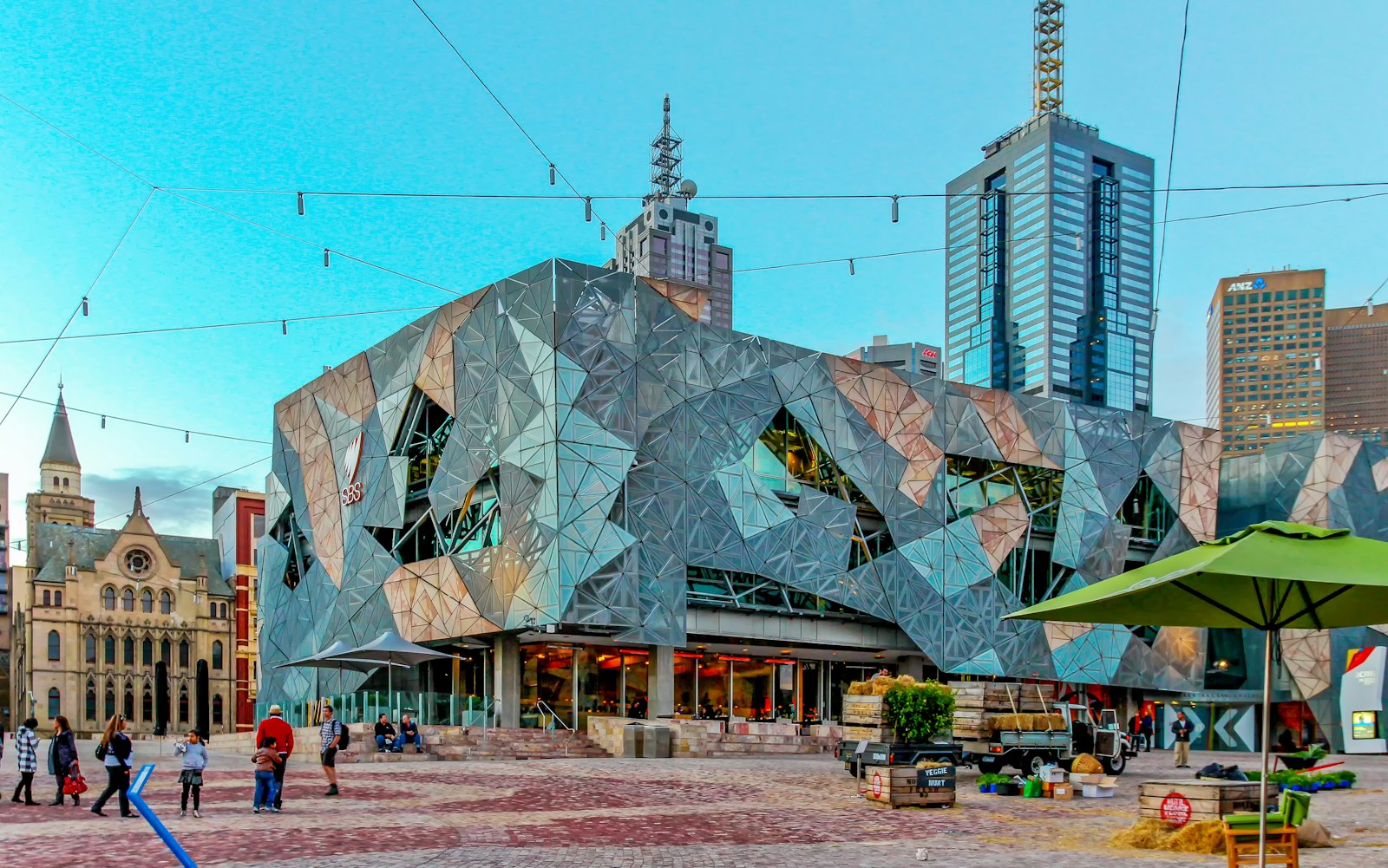 People crossing Federation Square beneath angular patterned façades, with Melbourne towers behind.