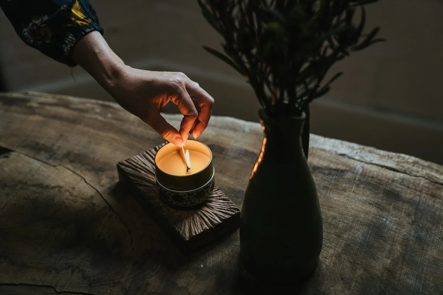 A hand lights a candle beside a dark vase on timber.