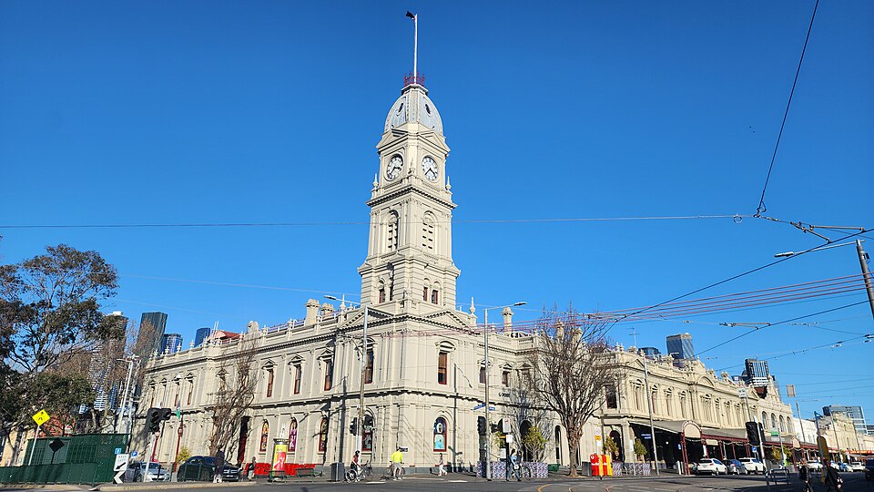 North Melbourne Town Hall’s pale façade and clock tower beneath tram wires and blue sky.